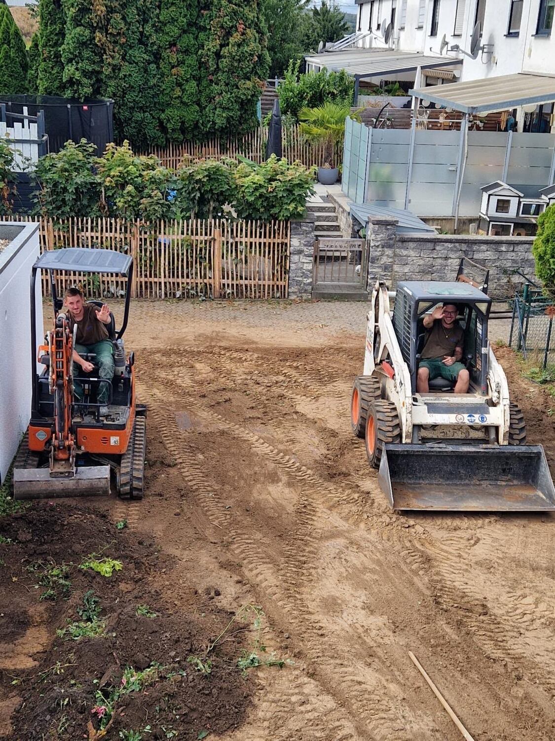 Zwei Mitarbeiter von Gies & Reitz winken aus Minibagger und Kompaktlader auf einer Gartenbaustelle